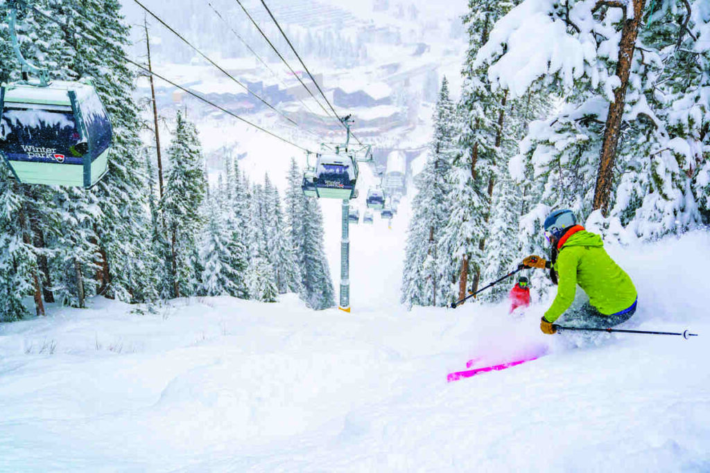 a skier with bright pink skis turns under a gondola line, through the trees, in deep fresh snow