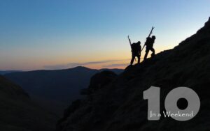 two skiers, skis on back, hike up mountain at dusk