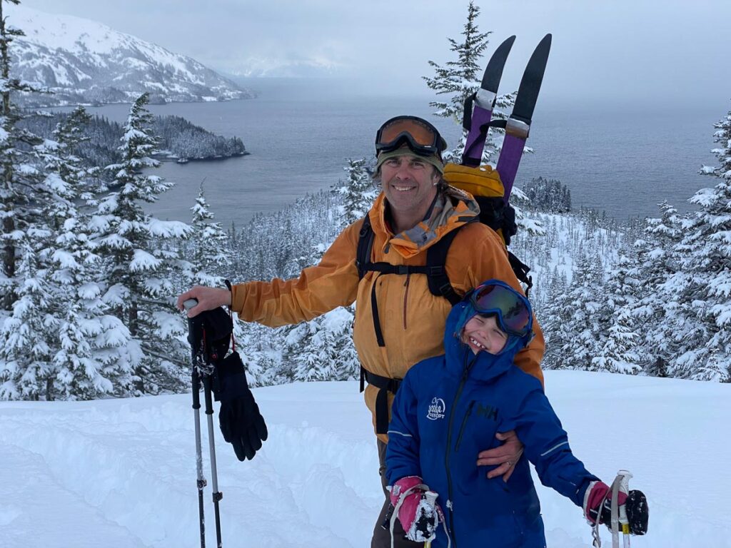man with skis on pack hugs young daughter as they stand on the mountain in deep, fresh snow, a sound of water below them