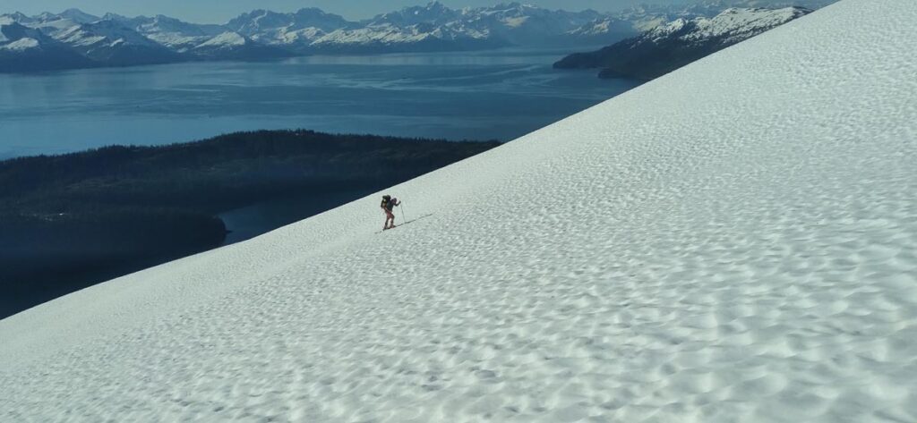a ski tourer heads up on steep sloping, pocked snow that is sun affected, a sound of water far below, as they summer ski in Alaska