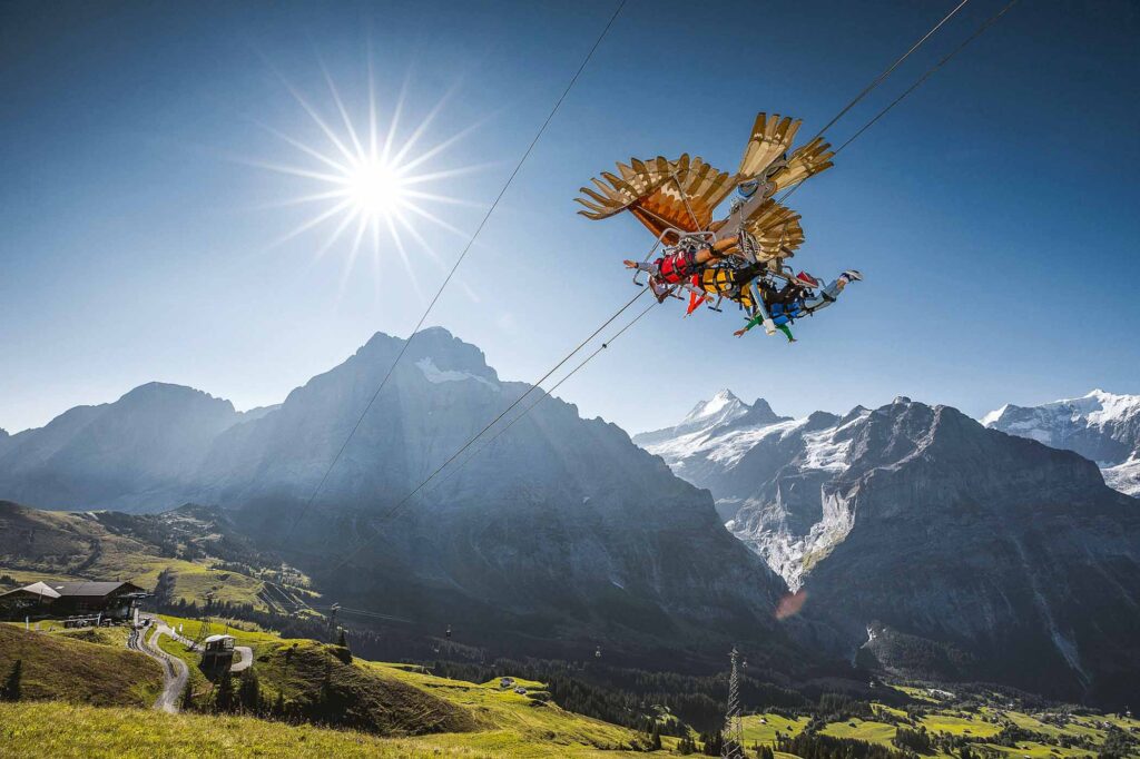 a golden bird aparatus on a zip line is photographed from below, with a human hanging underneath in some crazy zipline ride somewhere high in the mountains