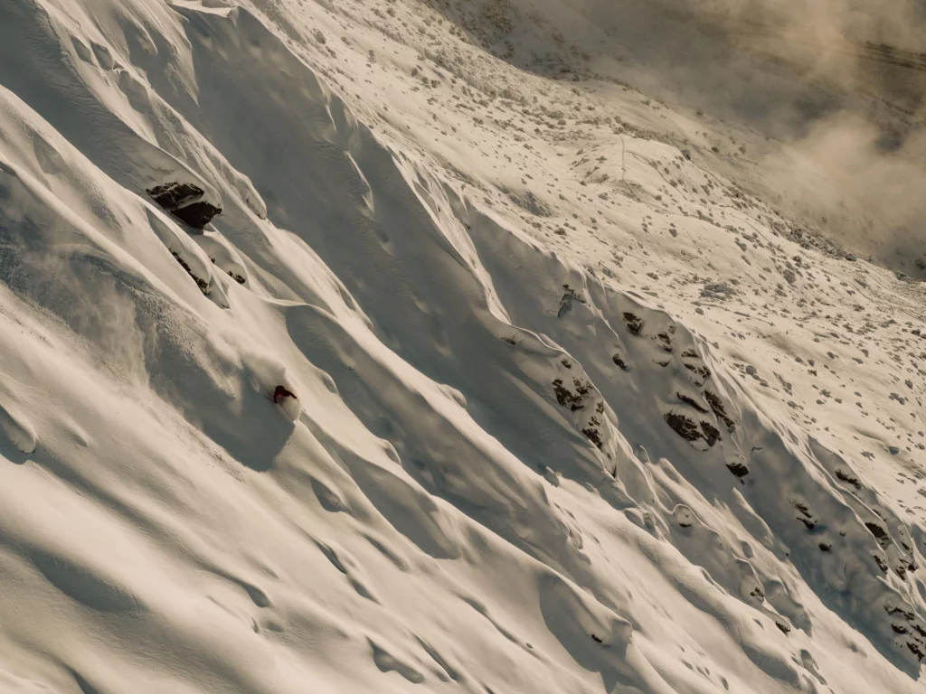 a skier on spines of a big, steep mountain, snow covering plenty of rock, under a yellowish, sepia light