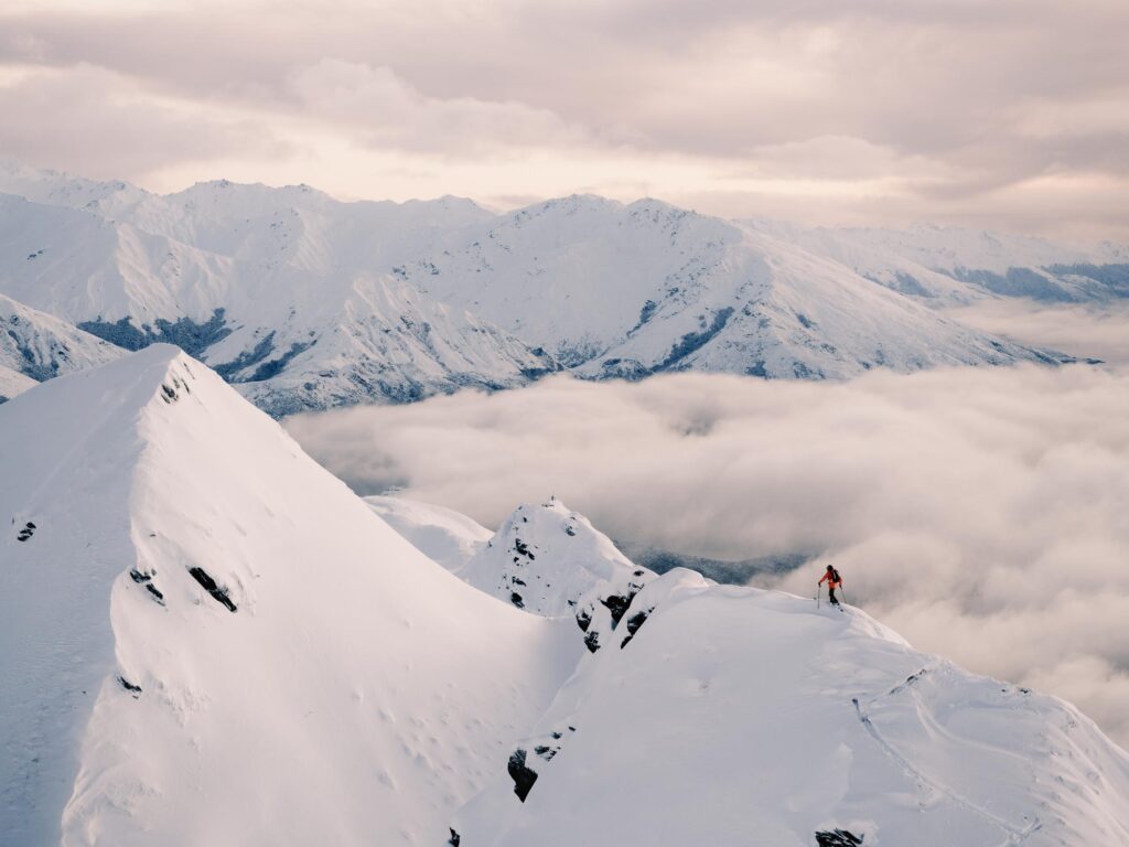 an aerial photo taken above the tops of mountains in New Zealand - snow covered mountains, cloudy sky, cloud filling the valley, and a skier in orange walking a ridgeline.