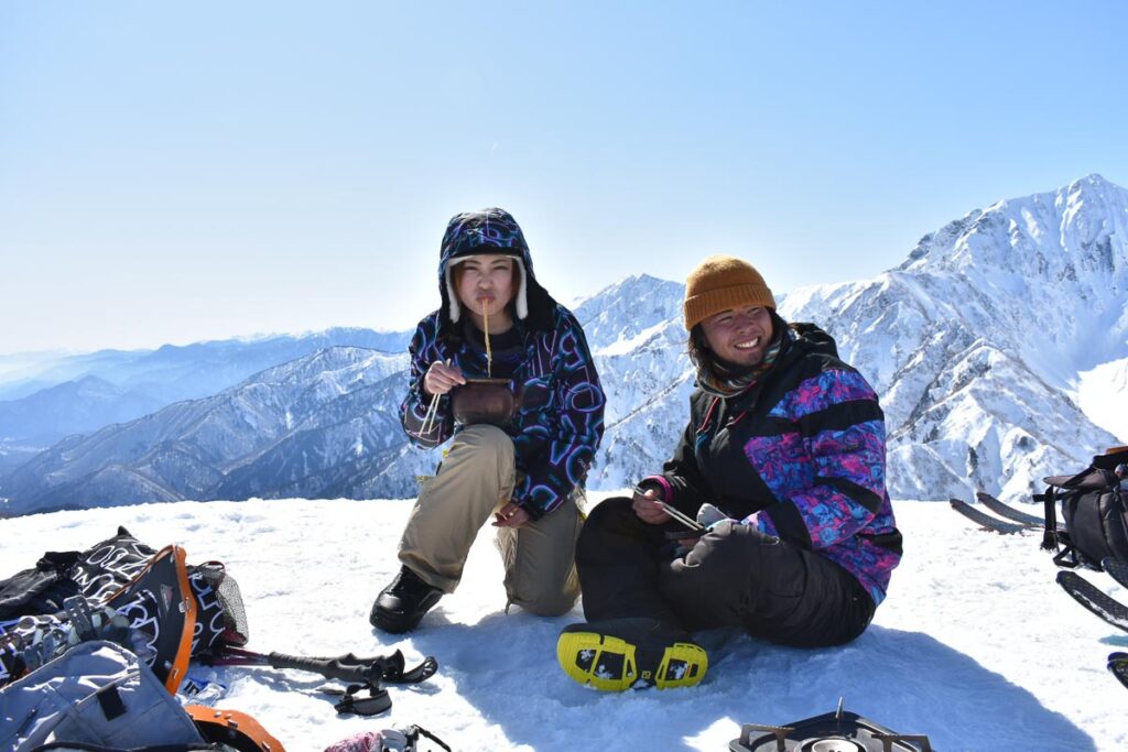 two Japanese splitboarders take a break on a snowy plateau having cooked up hot noodles on a camp stove, smiling and eating, with the snowy mountains behind (and even Mt Fuji visible in the background)