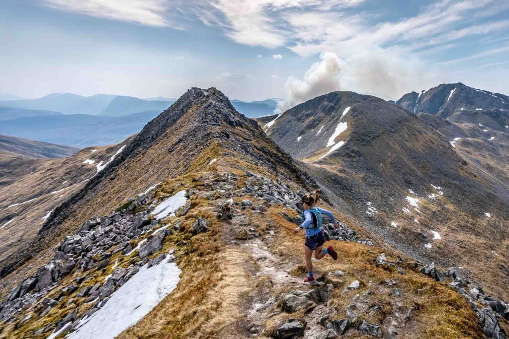 a woman running downhill on a rock trail path, patches of snow around in the mountain massif's peaks