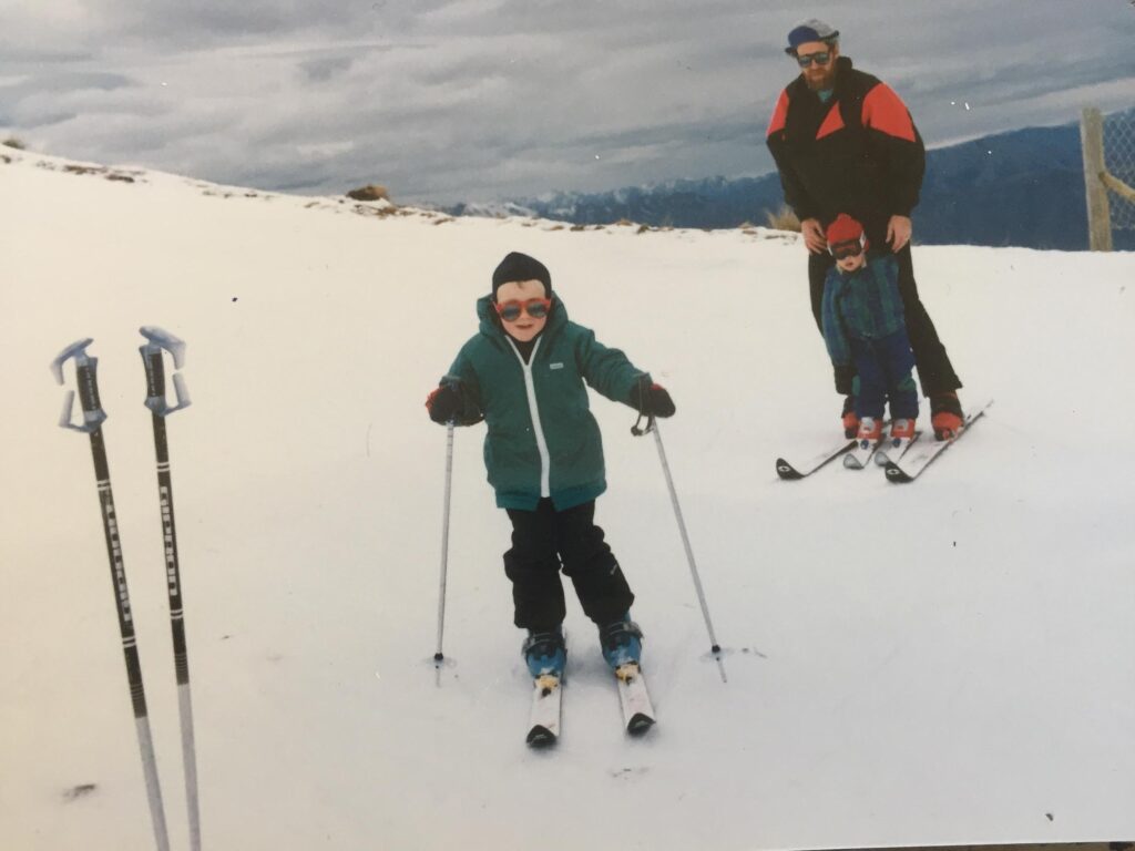 a child skiing in the 80s, this is pro skier Sam Smoothy, wearing red sunnies, another child between a father's skis behind, in an 80s photograph