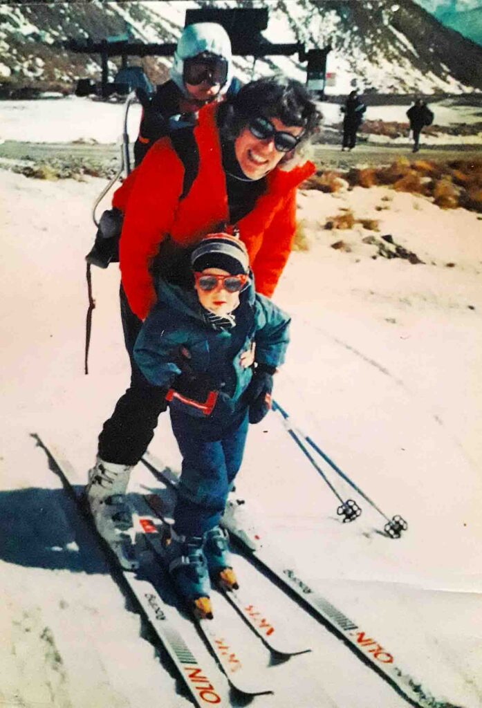 an 80s photograph of a young (4-year-old-ish) Sam Smoothy between his mum's skis (as she has another child in a pack on her back), smiling into the camera