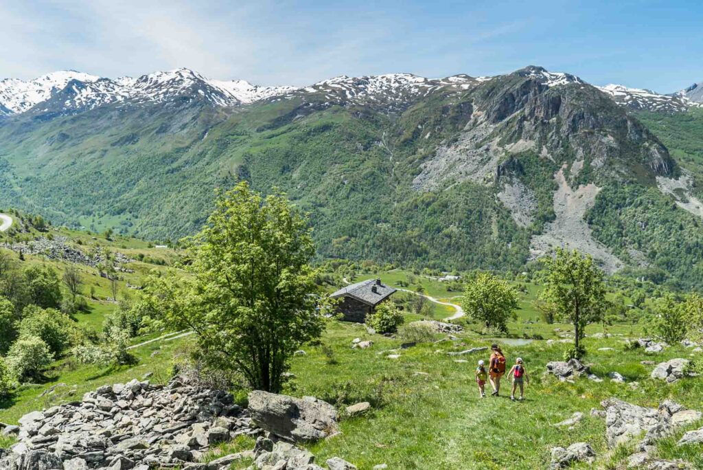 two kids and adult hike in high altitude mountains, surrounded by lush green grass with snow-capped mountains in the distance across a valley