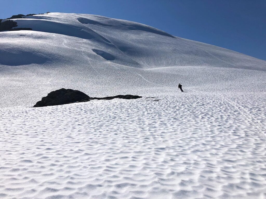 the pocked surface of  snow (from strong sun) is photographed with the silhouette of a single person summer skiing in Alaska