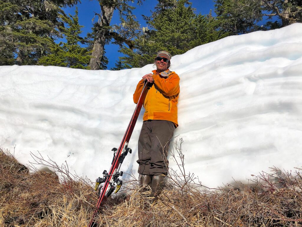man stands on snow, holding skis, against a wall of snow as tall as him