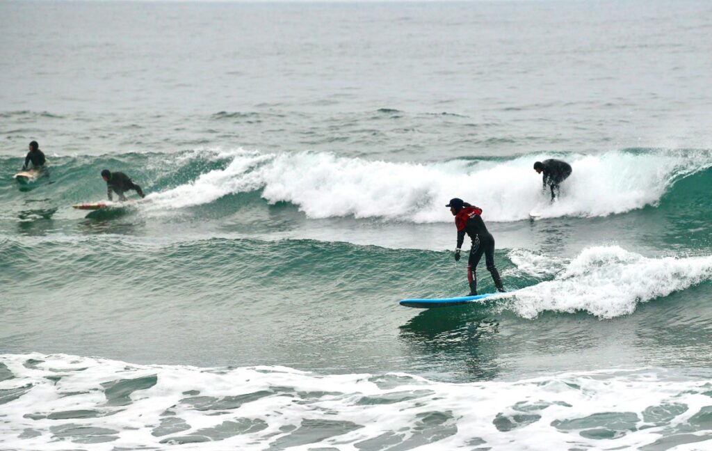 green sea on a moody day, with several surfers in bonnets and full wetsuits, surfing the modest waves