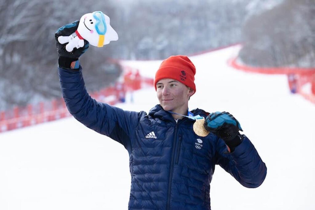 Young ski racer Zac Carrick-Smith holds up inflatable teddy, holding a gold medal, in celebration - as he stands on the snow