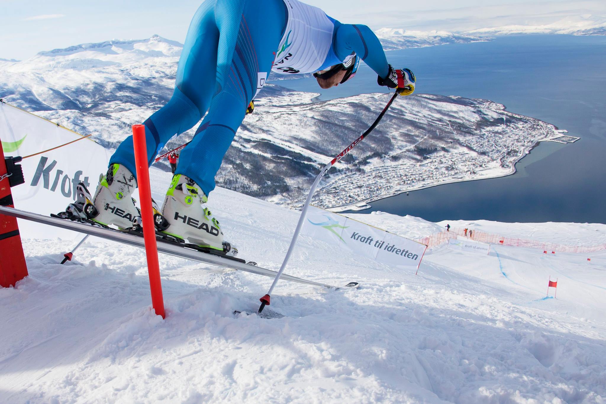 ski racer sets out of gates on a course above a fjord in what can only be Norway