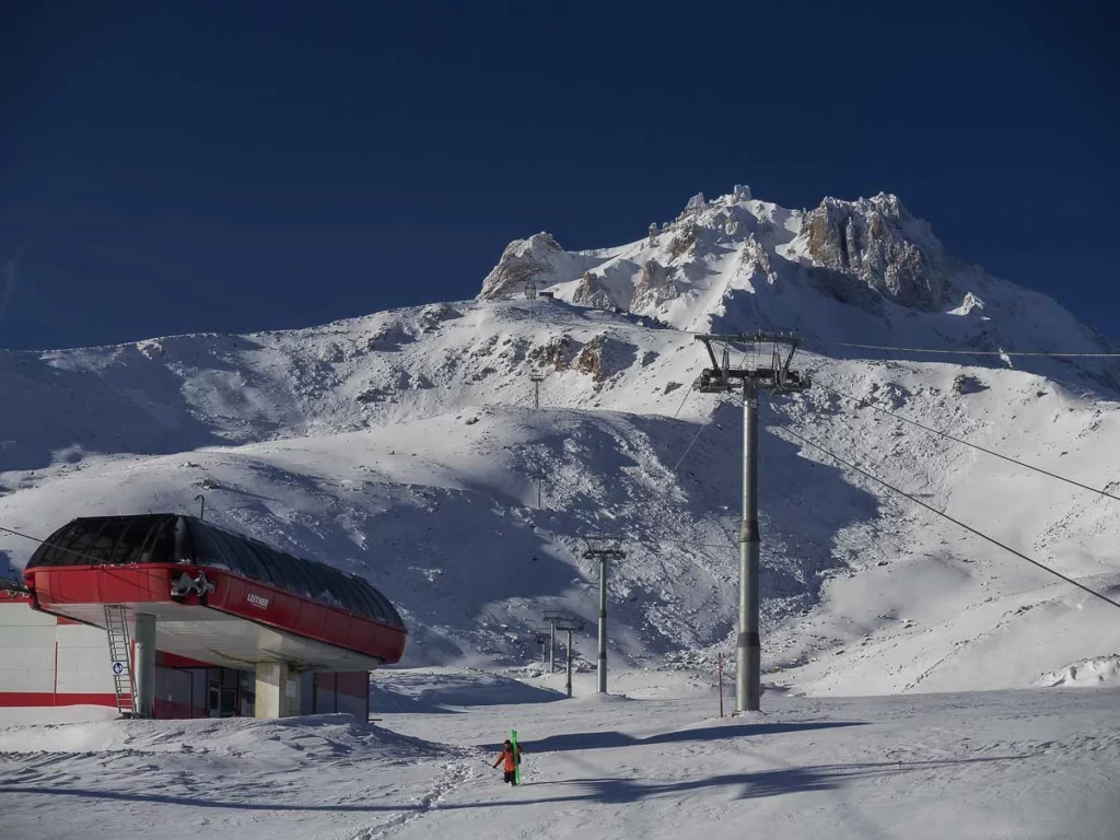 a red chairlift base station in Turkey, with a sole skier walking, skis in hand, towards camera