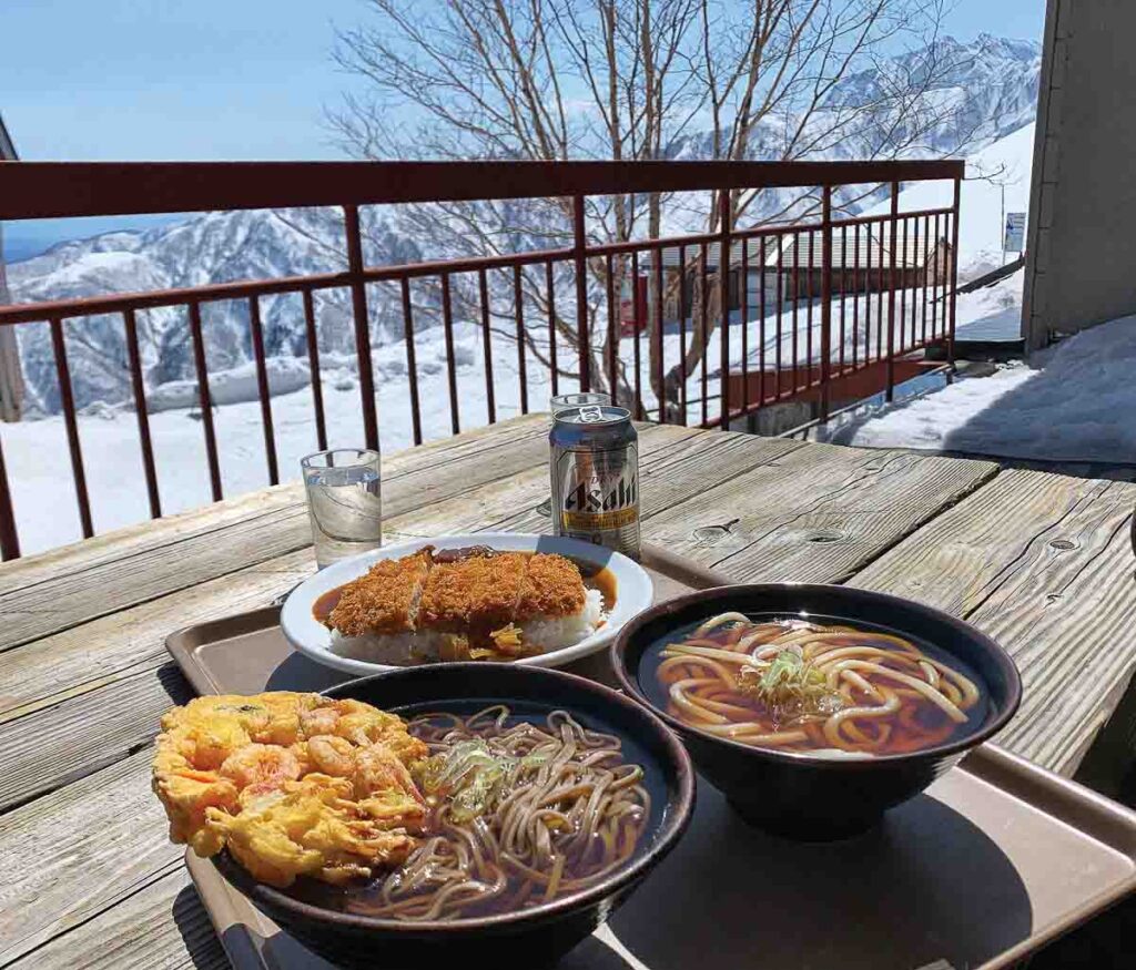 bowls of ramen, a katsu curry on a tray, and a Japanese Alps mountain backdrop