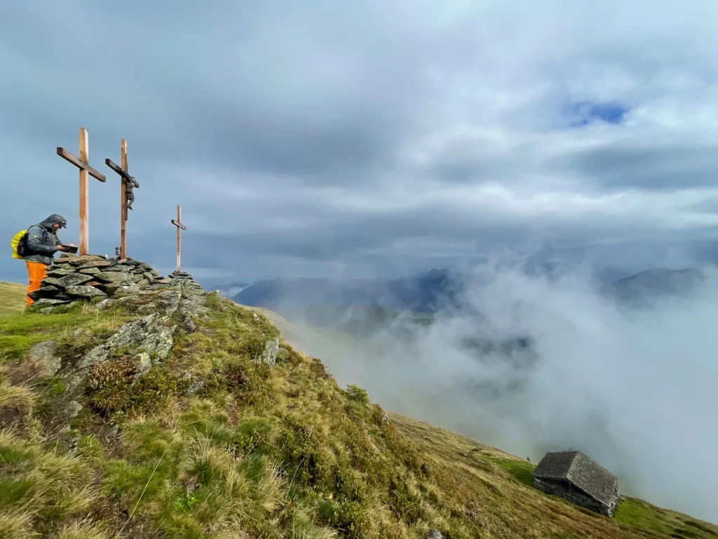 three crosses mark a mountain's peak, with a hiker standing behind one, looking over a valley on a cloudy day