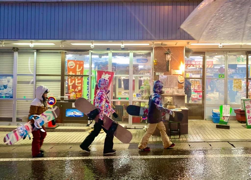 three snowboarders, hoods up, walk in front of a convenience store in the snow