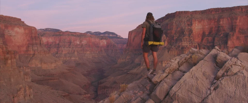 A man stands on a rock on the upper edges of the Grand Canyon, pink under an evening light