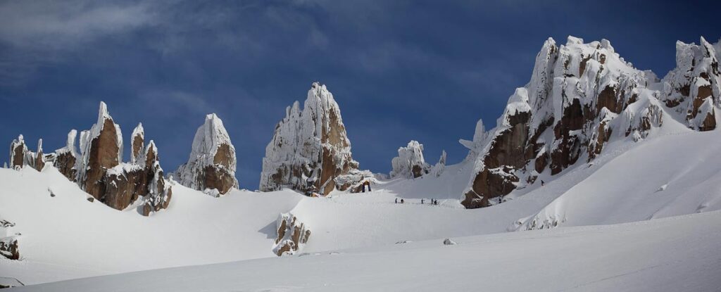 cathedral-spire like rocks in the high mountains, surrounded by snow and a collection of ant-sized skiers/mountaineers