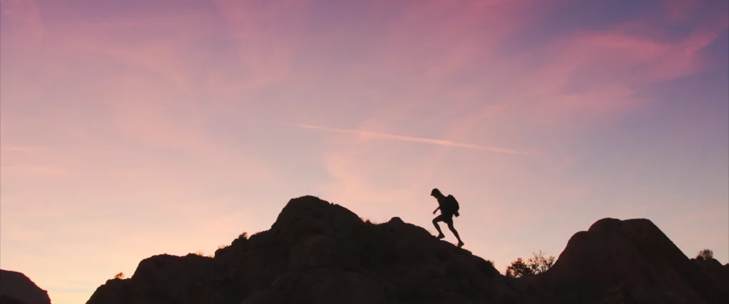 a silhouette of a person climbs over the ridge line of a rock, the sky glowing pink above at dusk