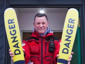 ski patroller in red jacket holds out skis (in front of patrol hut) that read danger