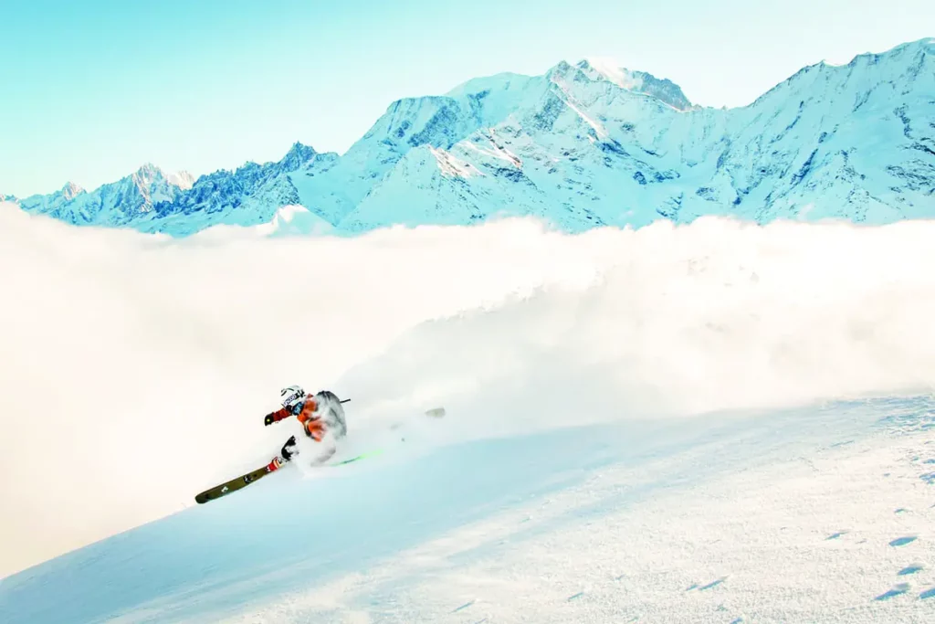 a telemark skier skis high in the mountains, judging by the surrounding peaks, looking over a valley filled with clouds, spraying snow behind them as they make a deep-leaning left turn