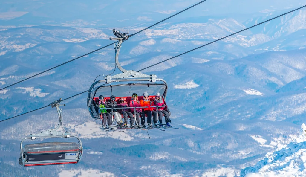 six sit on a chairlift, with just the forested hilly low-lands visible far below them in the background