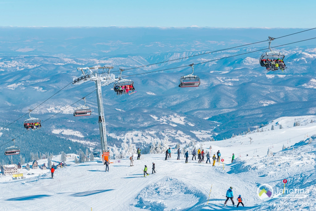 a busy ski hill is pictured, with skiers on a piste, a chairlift full of people travelling over their heads, and forested hilly lowlands far below