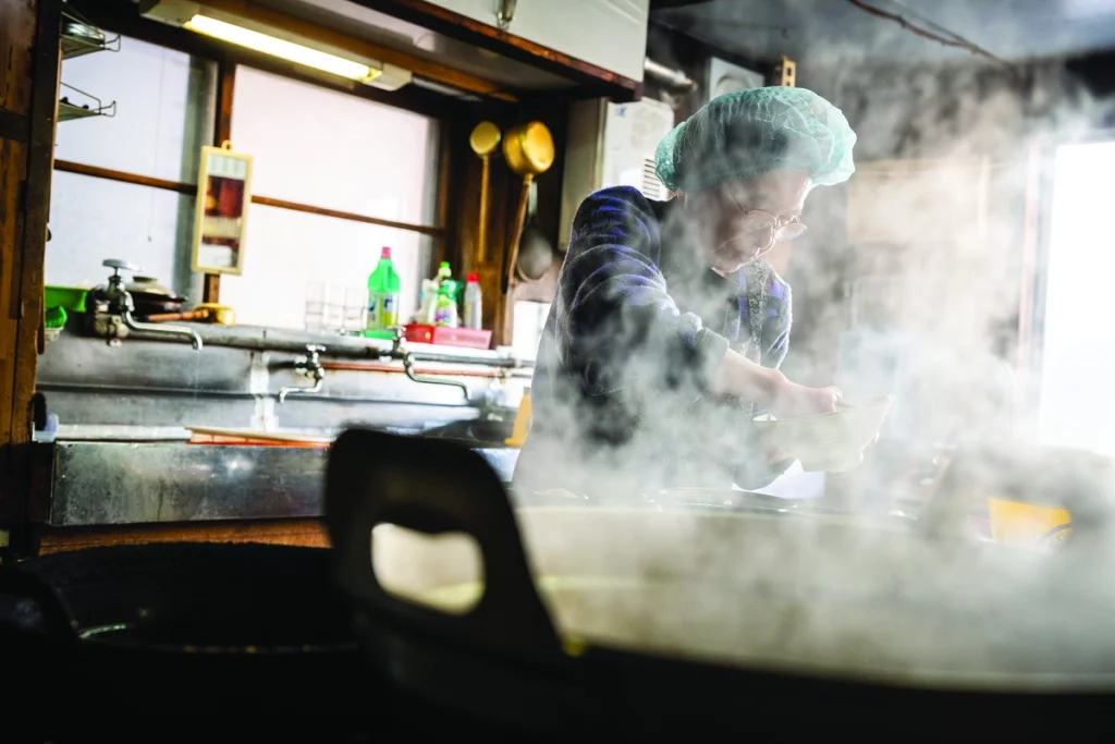 a Japanese woman in a hairnet is pictured cooking through the smoke of a pan in the foreground