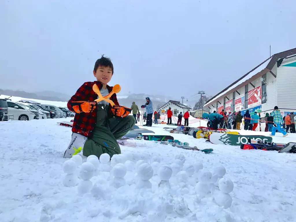 a boy smiles to camera at the base of a ski area, with an orange plastic snowball making device, and piles of snowballs ready made in front of him