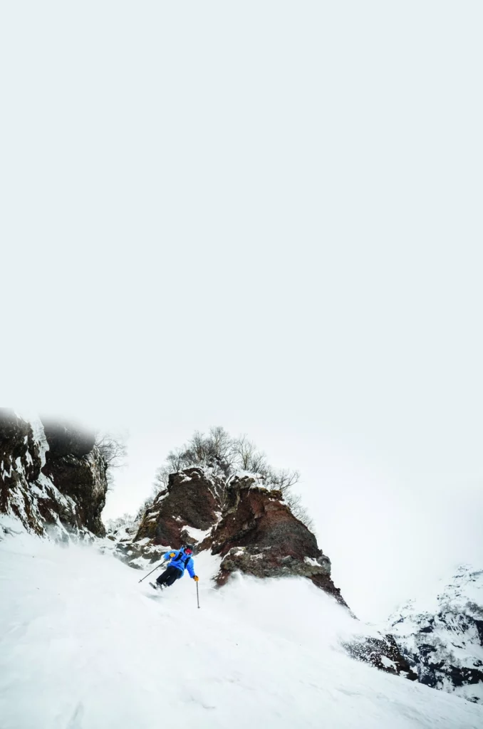 a skier in blue takes a little air on a turn, skiing off-piste by rocks on a day with no visibility