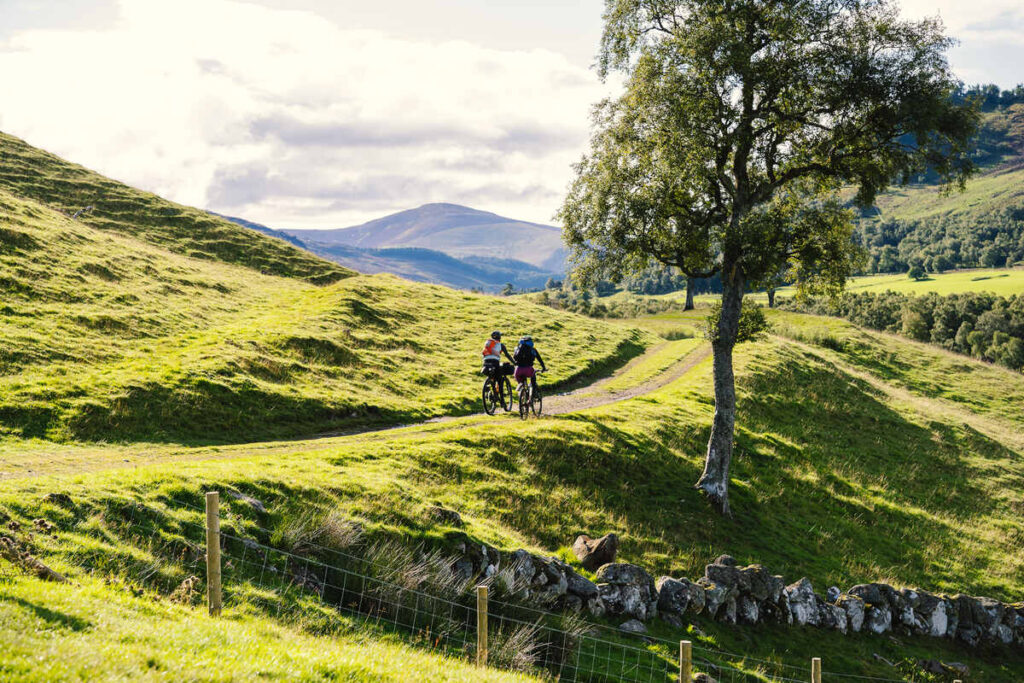 a lush, green landscape surrounded by rolling hills with two cyclists riding along on a dirt track