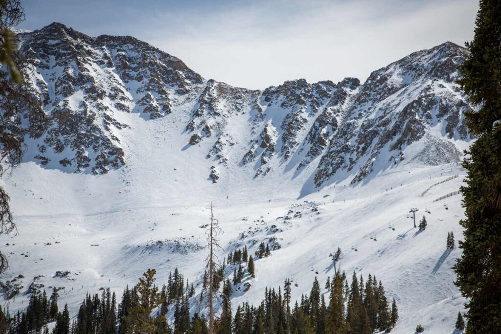 a mountain face is shown, covered in snow, skiers and lifts in front