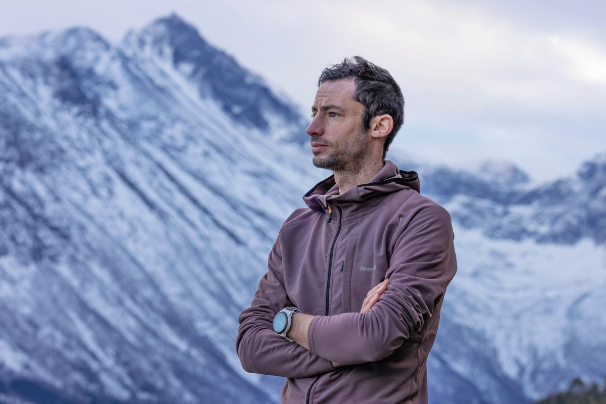 Ultra athlete Kilian Jornet stands in the high snowy mountains, arms crossed in a running jumper, looking to the side of the camera