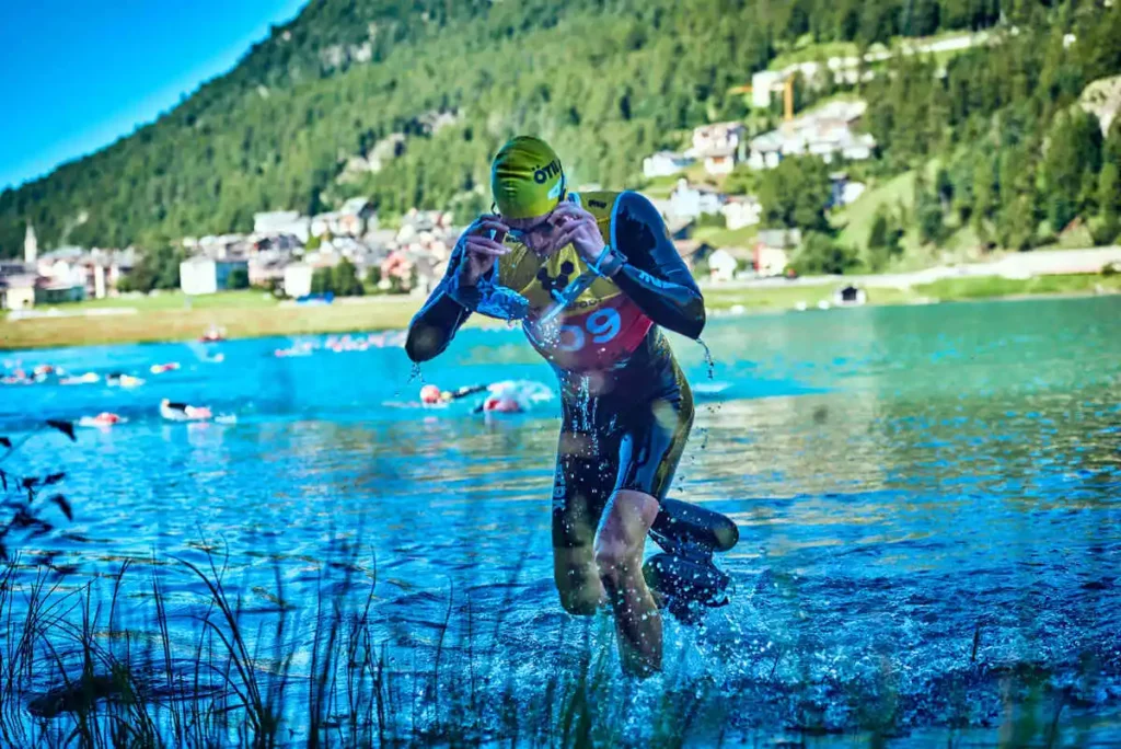 a sim-run swimmer steps out of the water, taking off goggles, by a blurred out mountain lake, having completed a swim section