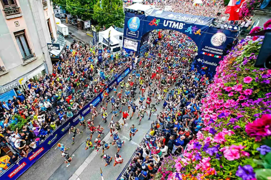 hundreds of runners set off through the starting gates of the UTMB in Chamonix, hundreds more spectators look on from the sidelines. The photo is taken from a birds-eye view from a balcony or something above